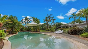 A swimming pool with a white umbrella, surrounded by palm trees and lounge chairs.