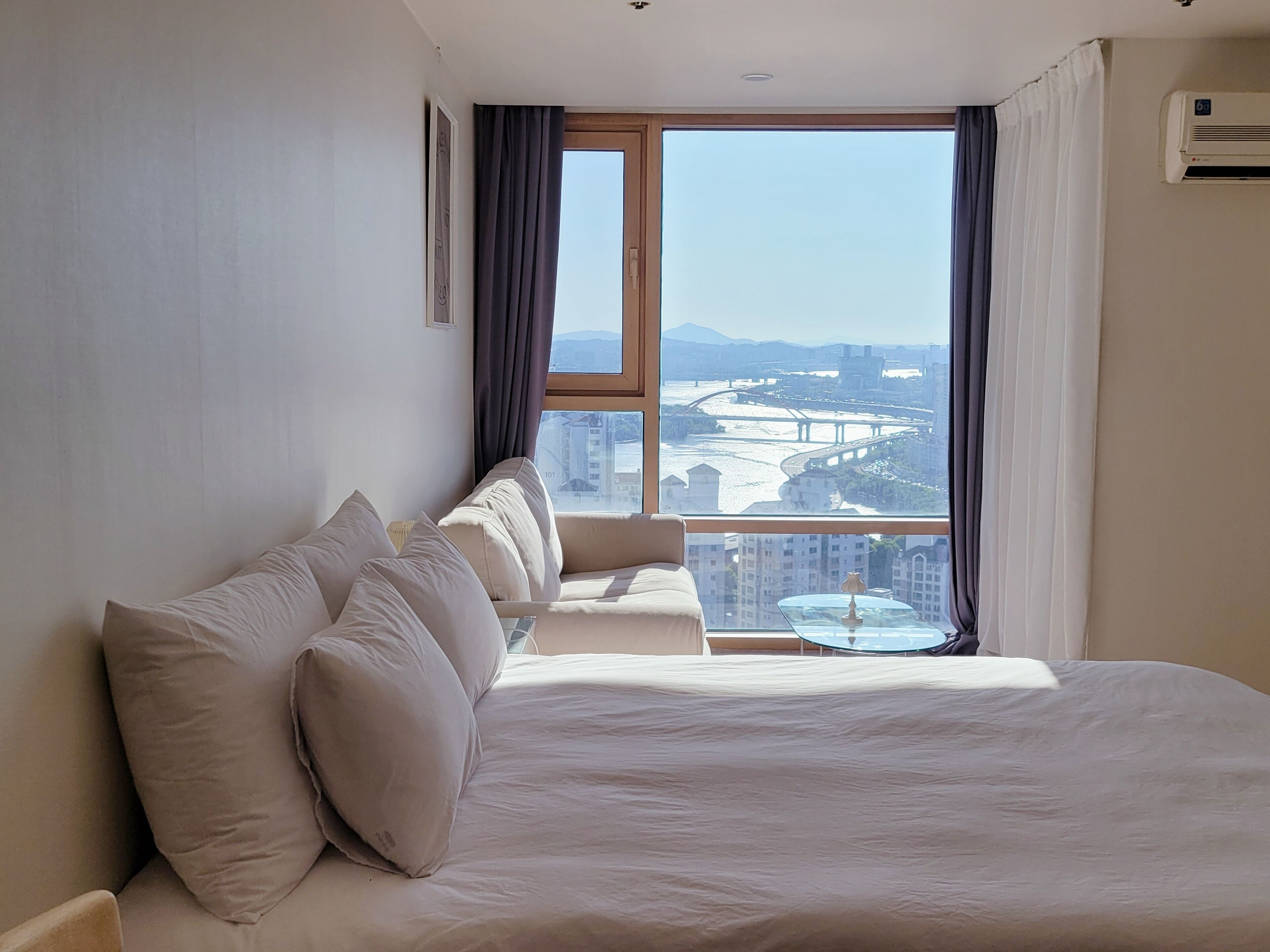 Habitación de hotel con una cama grande, vistas a un paisaje urbano y un equipo de aire acondicionado.
