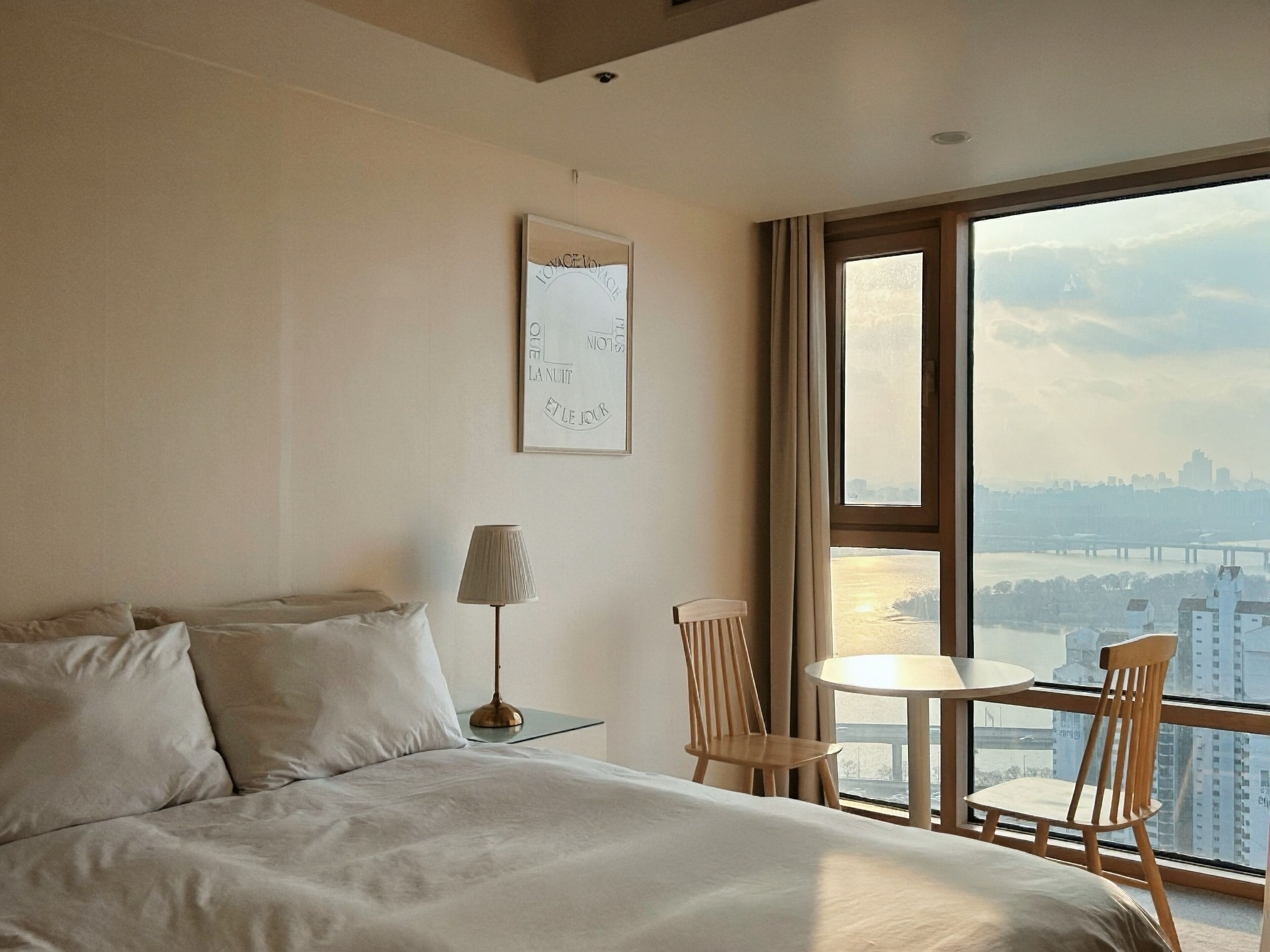 Habitación de hotel con cama, escritorio con lámpara, silla y vista panorámica de la ciudad a través de un ventanal amplio.