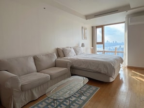 A modern hotel room with a large bed, a sofa, a glass coffee table, and a view of the city skyline through a large window.