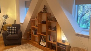 A cozy room with a sloped ceiling, a wooden bookshelf, a comfortable armchair, and a small table. - A brand new apartment decorated to a  high standard in a landmark development. (London)