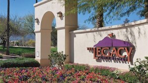Un letrero del Legacy Golf Resort, con un logo de paraguas morado, rodeado de flores y follaje coloridos.