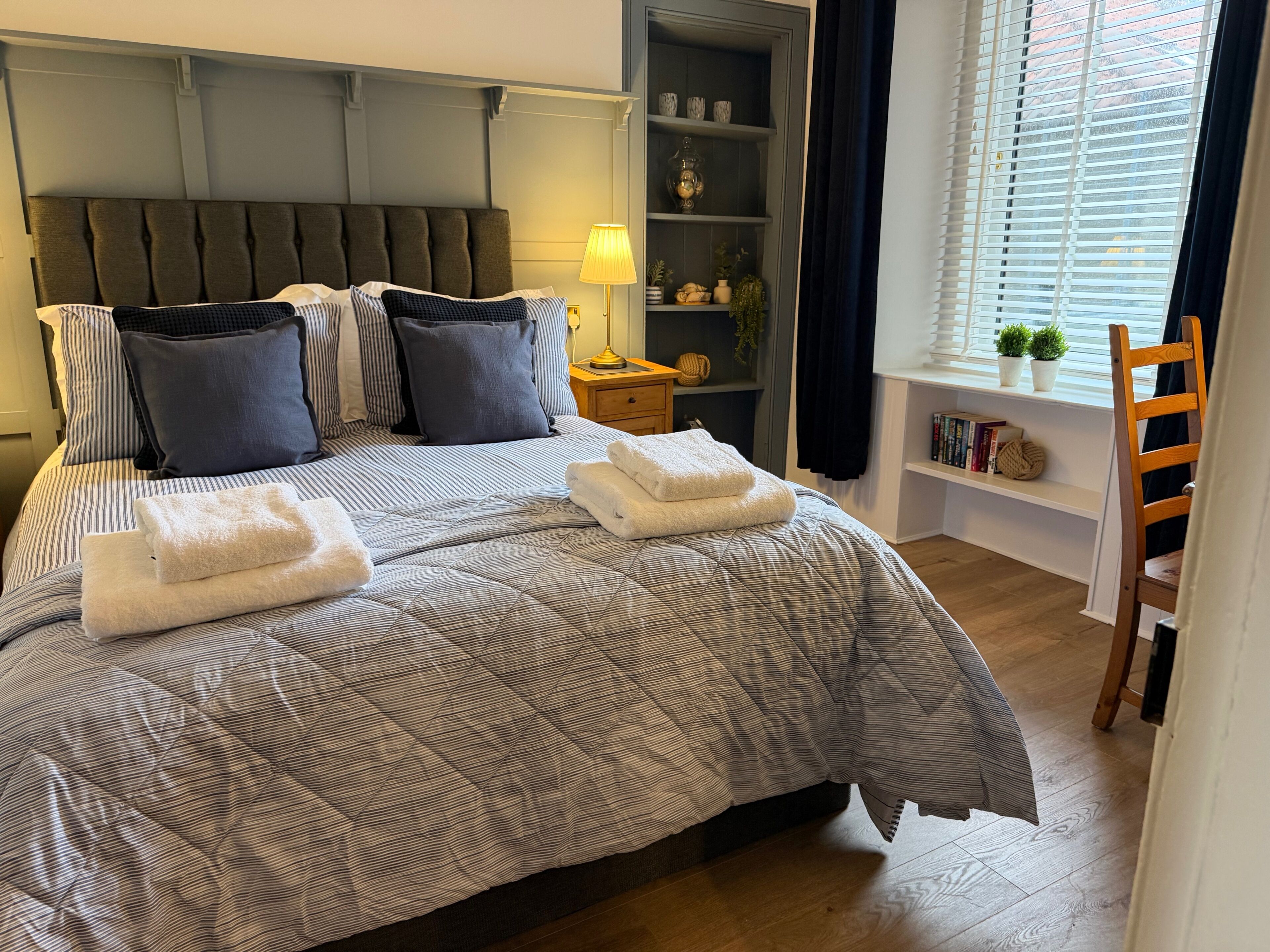 A neatly made bed with striped bedding, a bedside table with a lamp, and a window with blinds.