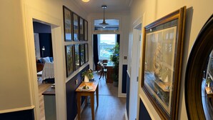 Ein Flur mit gerahmten Kunstwerken an den Wänden, ein Essbereich mit Tisch und Stühlen sowie ein Blick ins Freie durch ein Fenster.