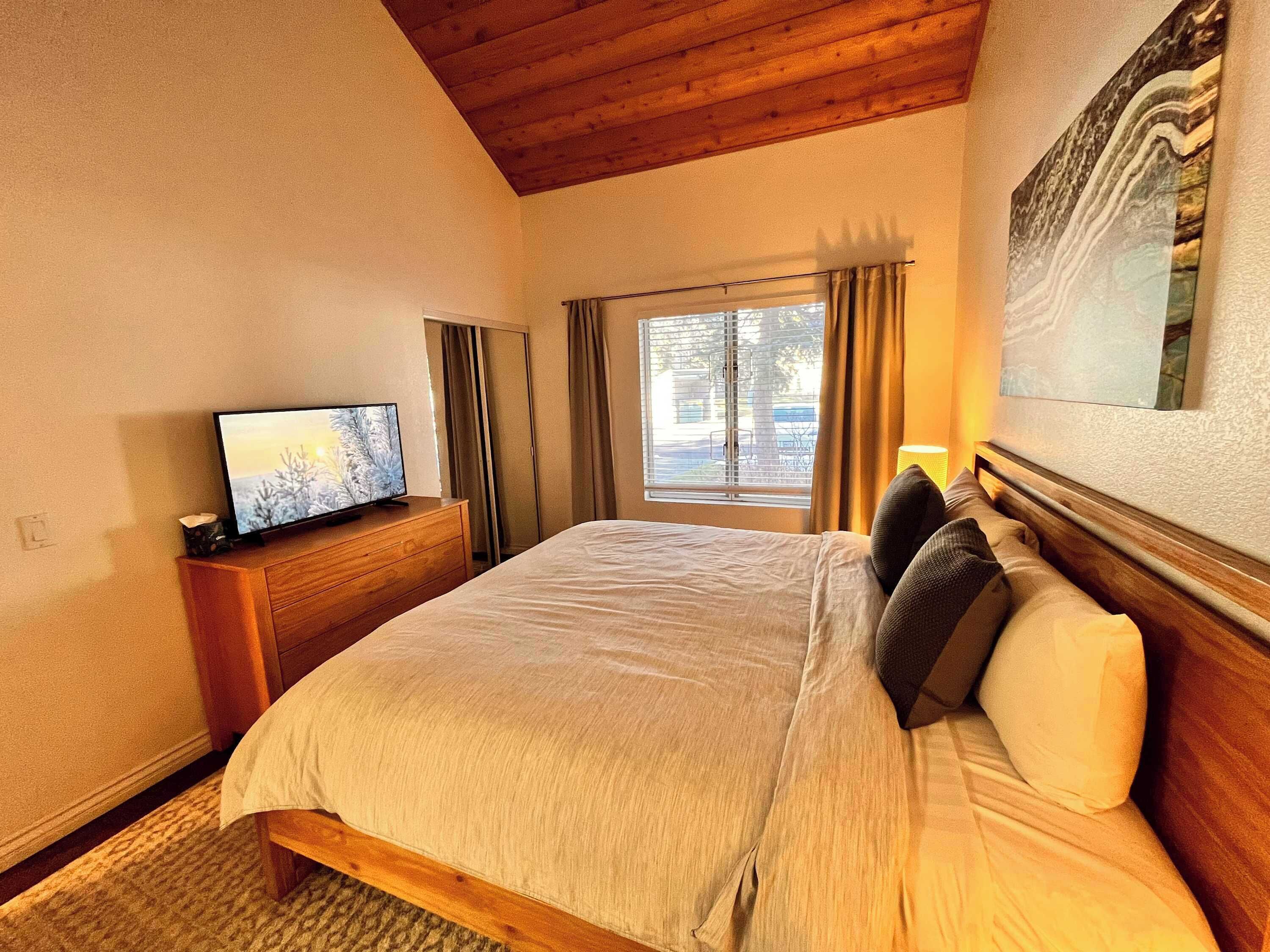 Une chambre d’hôtel avec un lit, un téléviseur posé sur une commode en bois, une fenêtre avec des rideaux et un tableau accroché au mur.
