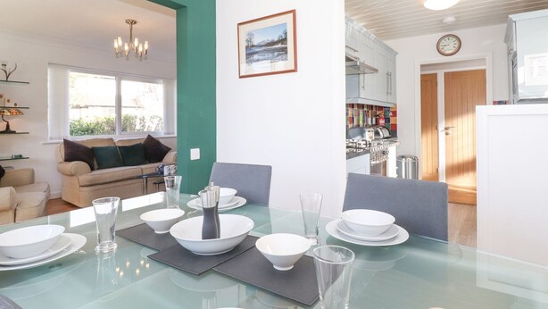 A modern kitchen with white cabinets, a dining table set with white plates and glasses, and a view into the living area with a sofa and a chandelier. - Wansfell View (Ambleside)