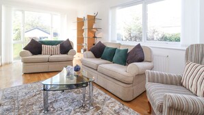 A spacious living room with two sofas, a glass coffee table, and a striped armchair. There is a chandelier, a bookshelf, and large windows providing natural light. - Wansfell View (Ambleside)