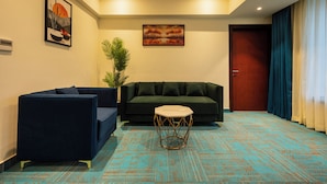 A modern living room with a dark blue sofa, a smaller armchair, and a geometric coffee table. There is a large painting above the sofa and a potted plant to the left. - Hotel Classic Pavilion (Thiruvananthapuram)
