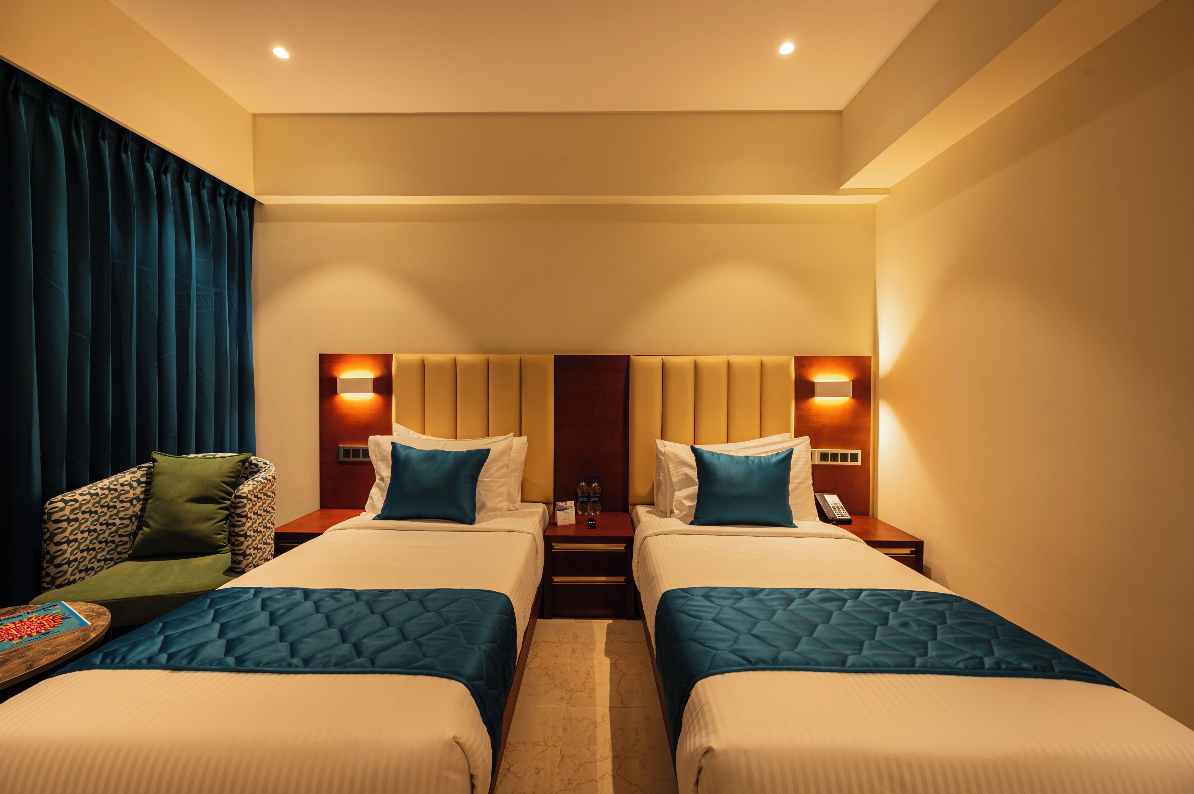 A hotel room with two beds, a nightstand with a phone and water bottles, a chair with a patterned cushion, and a wall-mounted lamp.