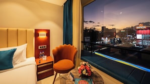 A hotel room with a bed, a chair, a nightstand, a table with a vase of flowers, and a large window with a city view. - Hotel Classic Pavilion (Thiruvananthapuram)