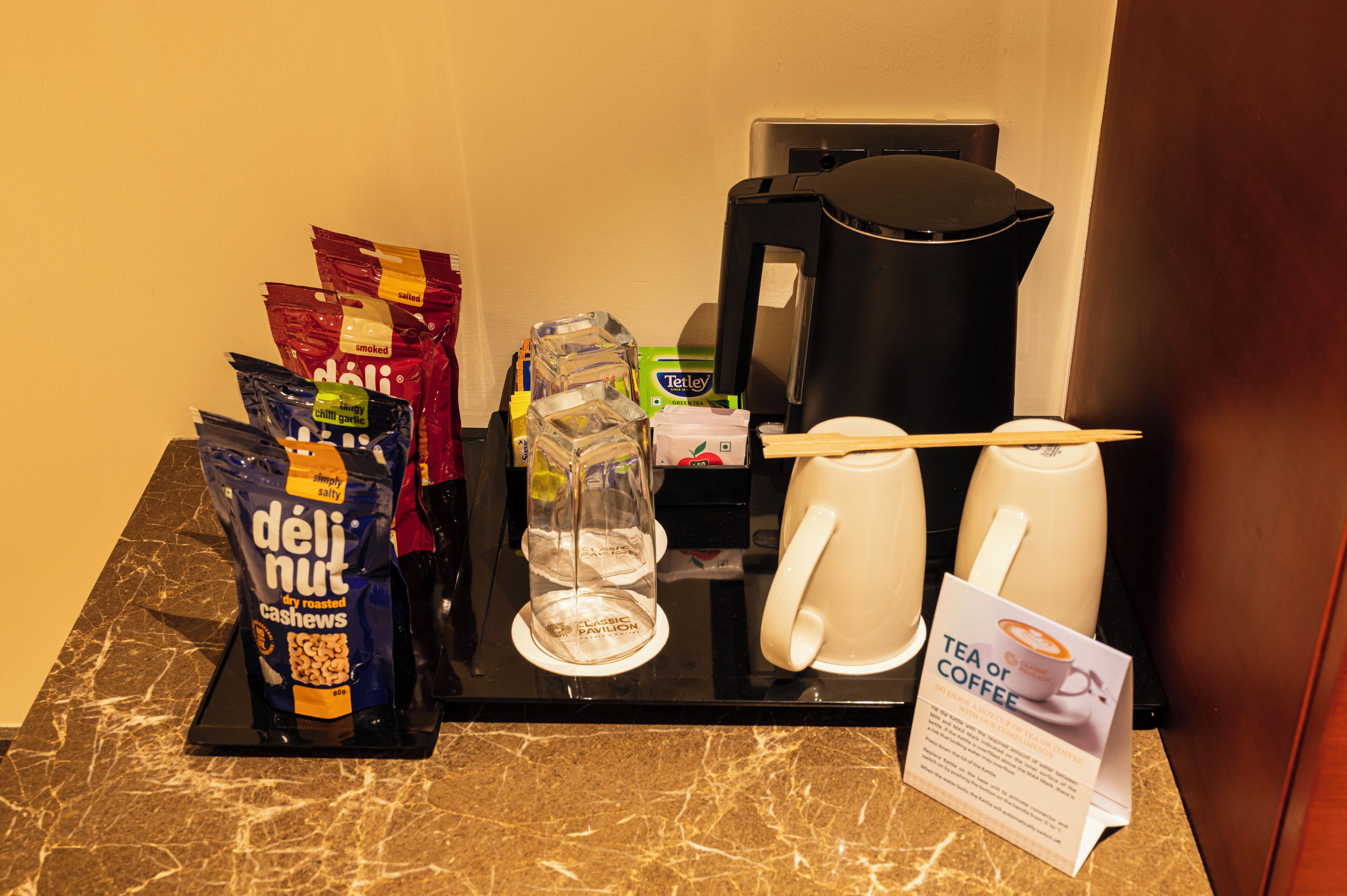 Una estación de café con cafetera, tazas, un letrero que indica té o café, y bolsas de bocadillos.