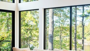 A modern living room with a beige sofa, a wooden coffee table, and large windows offering a view of trees. - Top Rated Muskoka - Sauna • Fireplace • Chef Kitchen (Gravenhurst)