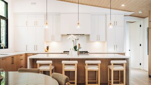 A modern kitchen with white cabinetry, a central island with bar stools, pendant lighting, and a visible orchid plant. - Top Rated Muskoka - Sauna • Fireplace • Chef Kitchen (Gravenhurst)
