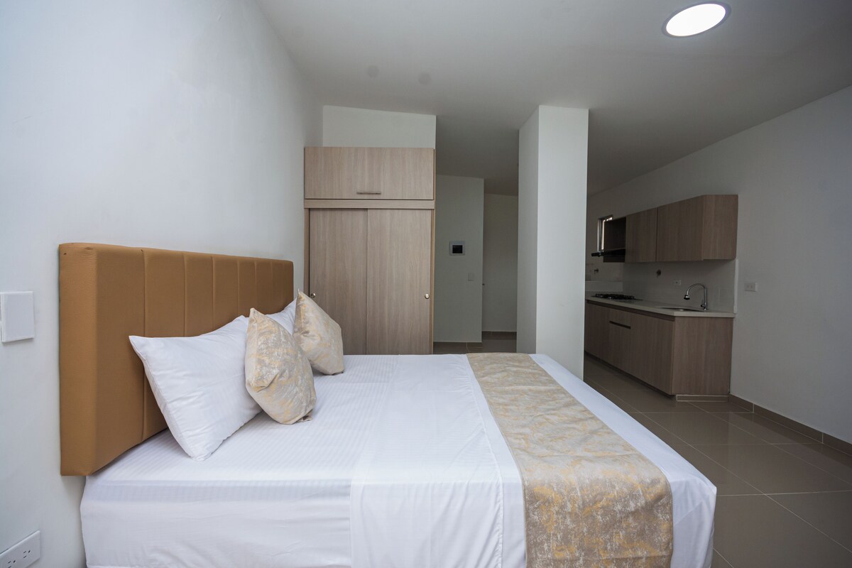 A hotel room with a bed, a wooden headboard, a kitchen area, and a bathroom visible through an open door.