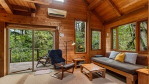 A wooden cabin interior with a sofa, a rocking chair, a small table, and a balcony with a view of the forest.