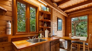 A wooden cabin interior with a kitchen area, wooden shelves, and a dining table.