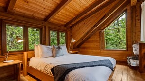 A wooden cabin bedroom with a bed, bedside lamps, and a view of the forest.