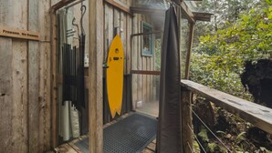 A wooden cabin with a surfboard, a sign reading 'Propane. No flames.', and a window with a view of the forest.