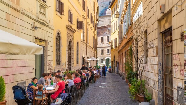 Rua de paralelepípedos estreita, ladeada por edifícios, com áreas de estar ao ar livre para cafés.