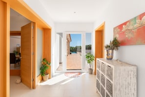A hallway with a wooden door, potted plants, a white console table, and a view to an outdoor area with a pool.
