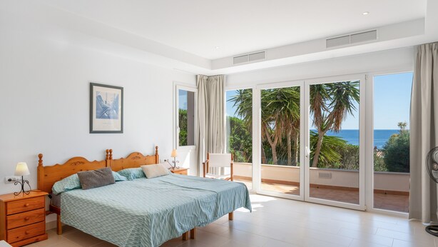 Een slaapkamer met een bed, nachtkastjes, een stoel en een grote schuifglazen deur die leidt naar een balkon met uitzicht op palmbomen en de zee.