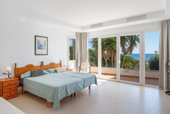 A bedroom with a bed, bedside tables, a chair, and a large sliding glass door leading to a balcony with a view of palm trees and the sea.