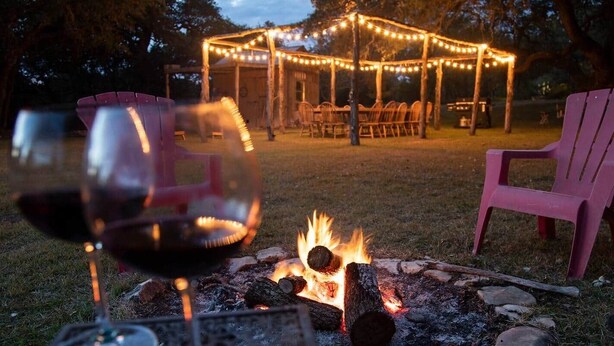 Een kampvuur met houtblokken en een glas rode wijn op de voorgrond, een houten pergola met lichtjes in de achtergrond, en een roze stoel aan de rechterkant.