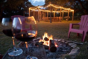 Ein Lagerfeuer mit Holzscheiten im Vordergrund, ein Glas Rotwein, im Hintergrund ein Holzkiosk mit Lichterketten und ein rosa Stuhl auf der rechten Seite.