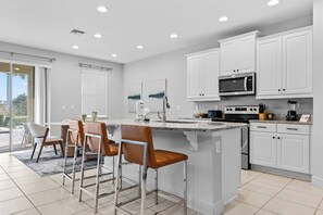 A modern kitchen with white cabinets, a central island with bar stools, and a dining area with chairs. - 4BR house with pool table near Disney (Kissimmee)