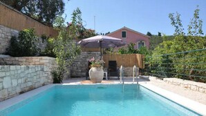 A rectangular swimming pool with a stone wall on one side and a wooden fence on the other. There is a chair and a large vase with flowers near the pool's edge. - Villa Adamantia in Ozias (Paxos)
