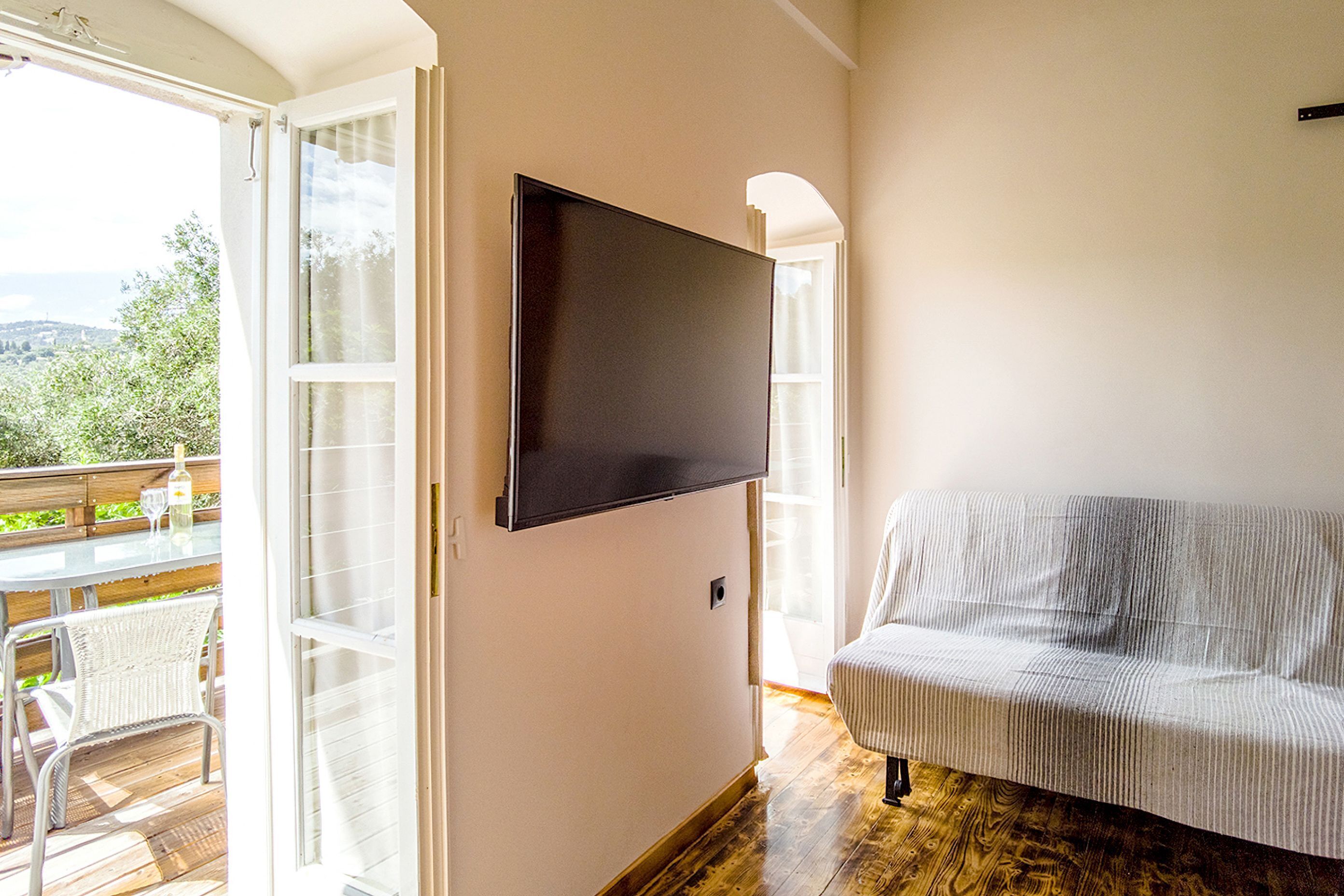 A room with a large flat-screen TV mounted on the wall, a white sofa, and a sliding glass door leading to an outdoor deck with a table and chairs.