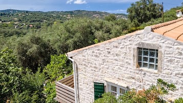 A stone building with a window and a wooden deck, surrounded by greenery and hills.