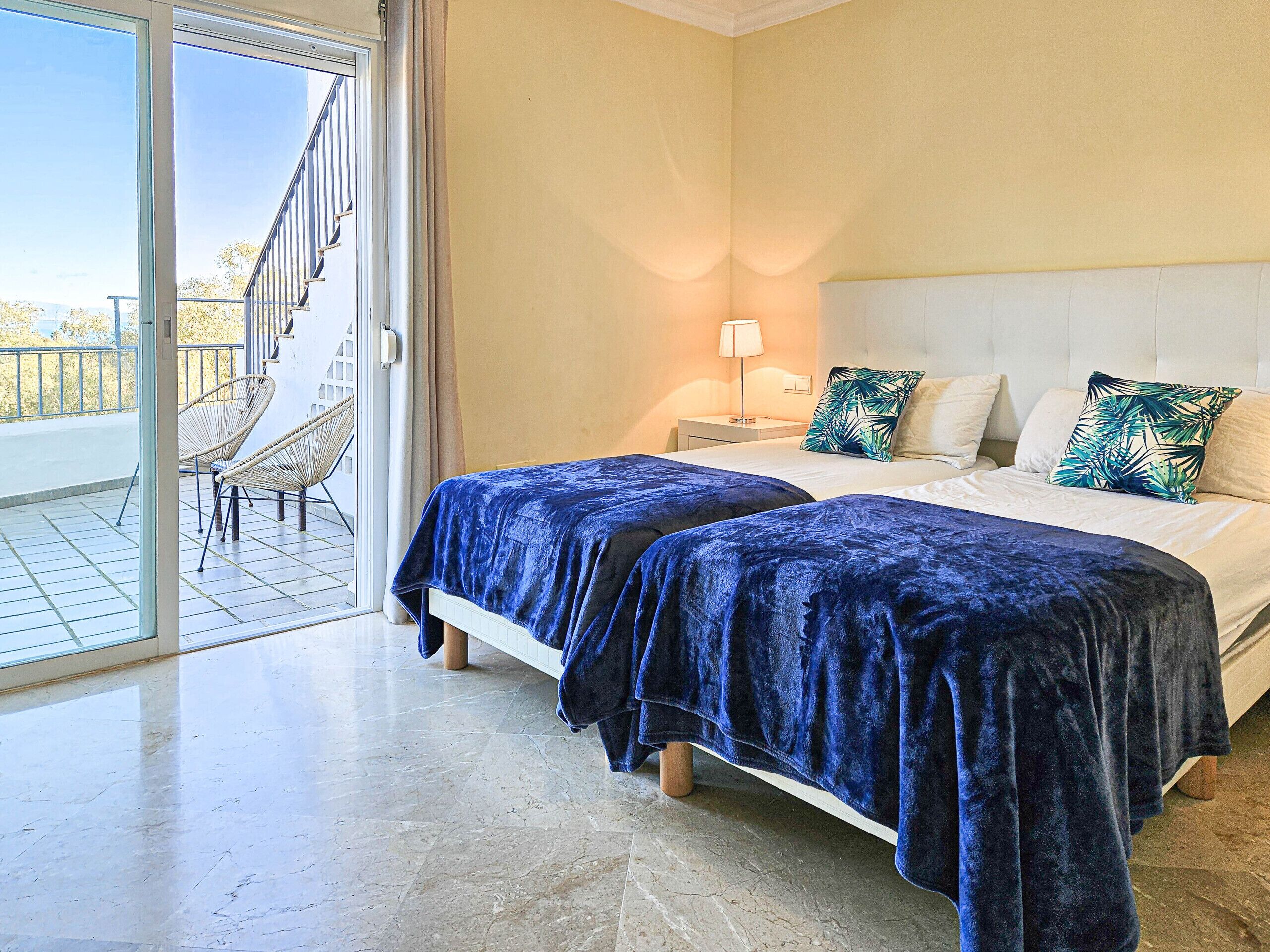 Ein Hotelzimmer mit zwei Betten, einem Balkon mit Korbstühlen und Blick auf ein Gebäude.