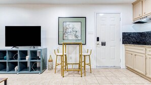 Una sala de estar con televisor, una obra de arte enmarcada, una puerta blanca y una zona de cocina con armarios.
