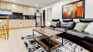 Cocina moderna con armarios de madera clara, un refrigerador de acero inoxidable y un microondas. Hay una alfombra blanco y negro en el suelo, y un sofá de cuero negro con cojines en la sala.