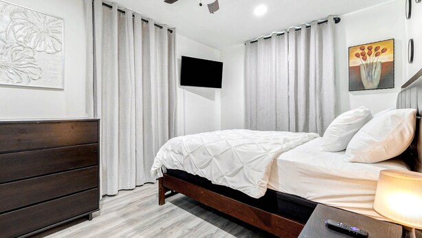 Un dormitorio con cama, cómoda de madera, televisor montado en la pared y lámpara en la mesita de noche.
