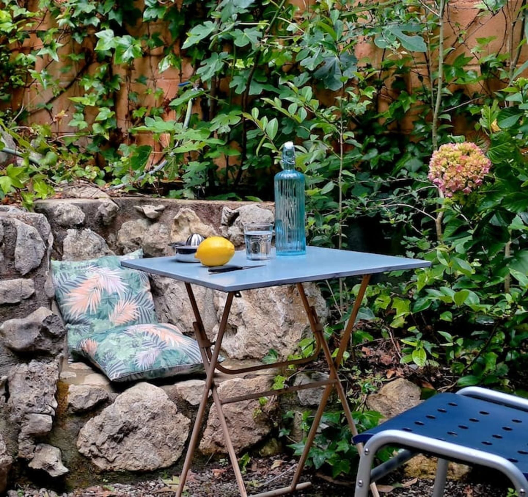 Un piccolo tavolo all'aperto con una bottiglia, un limone e un bicchiere, addossato a un muro in pietra e circondato da vegetazione rigogliosa.