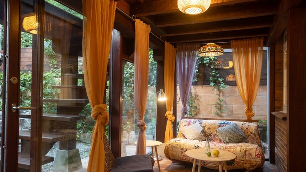 Una habitación acogedora con sofá, una mesita y una silla. Se ve una ventana con cortinas y vistas a la vegetación.