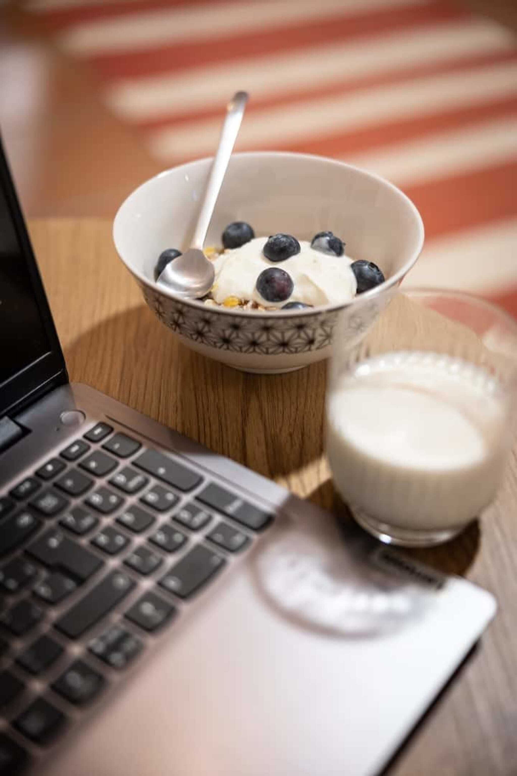 Ein Laptop auf einem Holztisch zusammen mit einer Schüssel Müsli und einem Glas Milch.