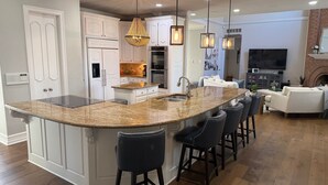 A spacious kitchen with a large island, bar stools, and a fireplace in the living area. - Luxury 4,300 SqFt Home • Sleeps 12 • Outdoor Kitchen & Patio with Large Pool (Colleyville)