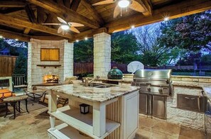 An outdoor kitchen with a stone fireplace, a grill, and a sink. - Luxury 4,300 SqFt Home • Sleeps 12 • Outdoor Kitchen & Patio with Large Pool (Colleyville)