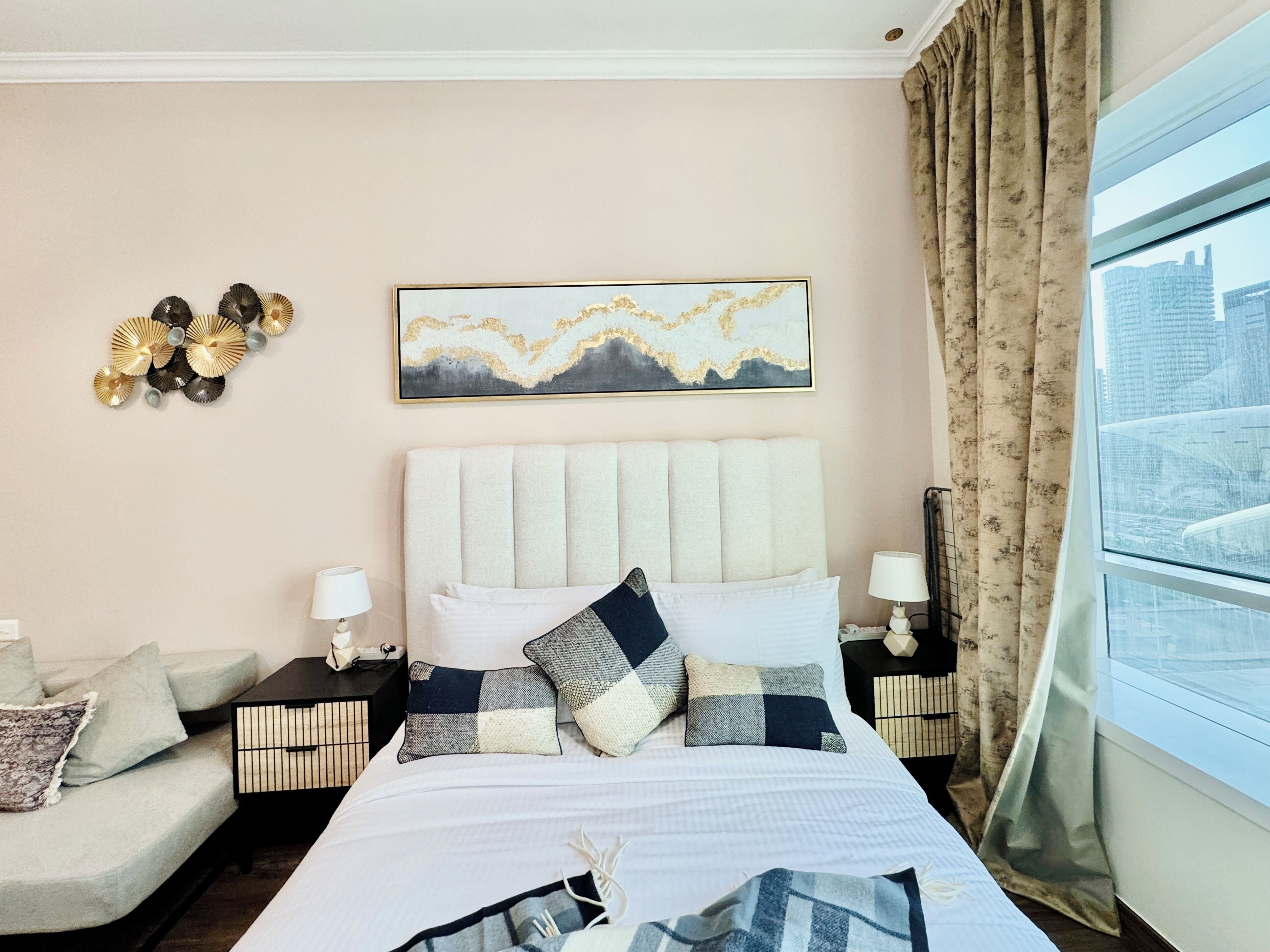 A hotel room with a large bed, a sofa, a decorative wall piece, and a window with a city view.