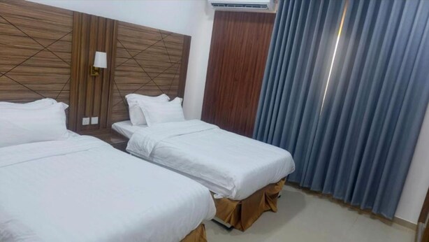 Habitación de hotel con dos camas individuales, cabecera de madera, lámpara de pared y ventana con cortinas azules.