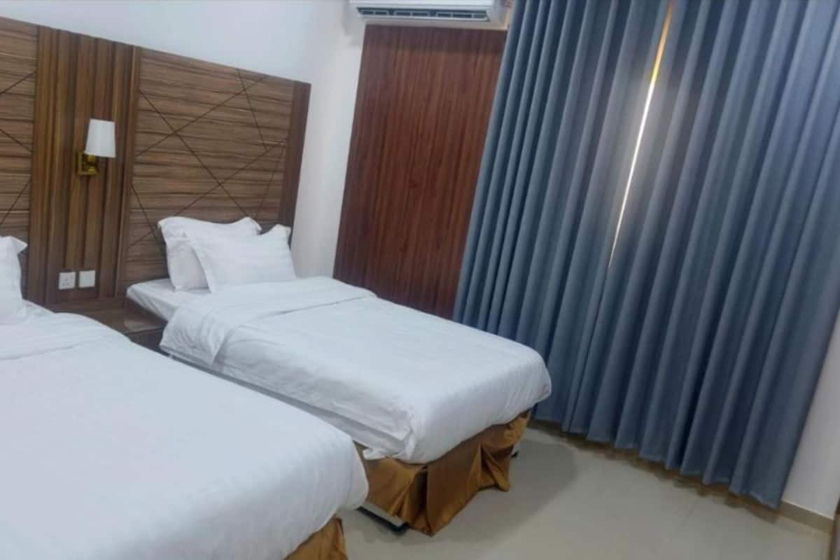 Habitación de hotel con dos camas individuales, cabecera de madera, lámpara de pared y ventana con cortinas azules.