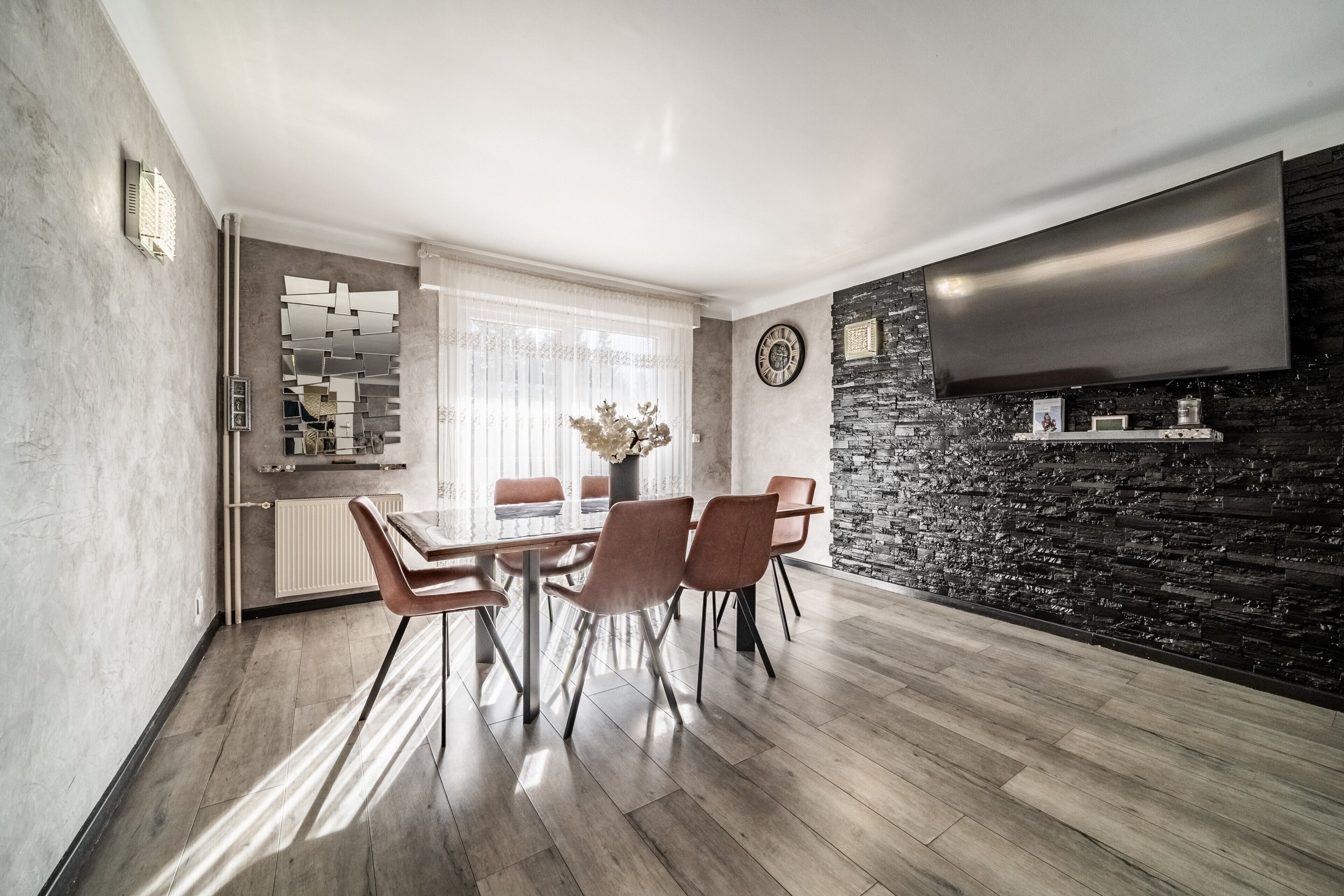 A modern dining area with a wooden table and chairs, a stone accent wall, a flat-screen TV, and a kitchen in the background.