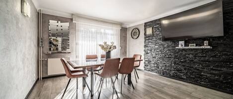 A modern dining area with a wooden table and chairs, a stone accent wall, a flat-screen TV, and a kitchen in the background.