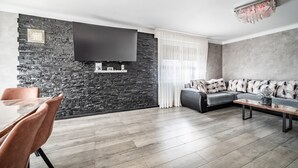 A modern living room with a stone wall, a flat-screen TV, a grey sofa, and a wooden coffee table. - Villa "L'Oranaise" – 190 m², Mountain View, Private Balconies, Terraces & Garden (Cluses)