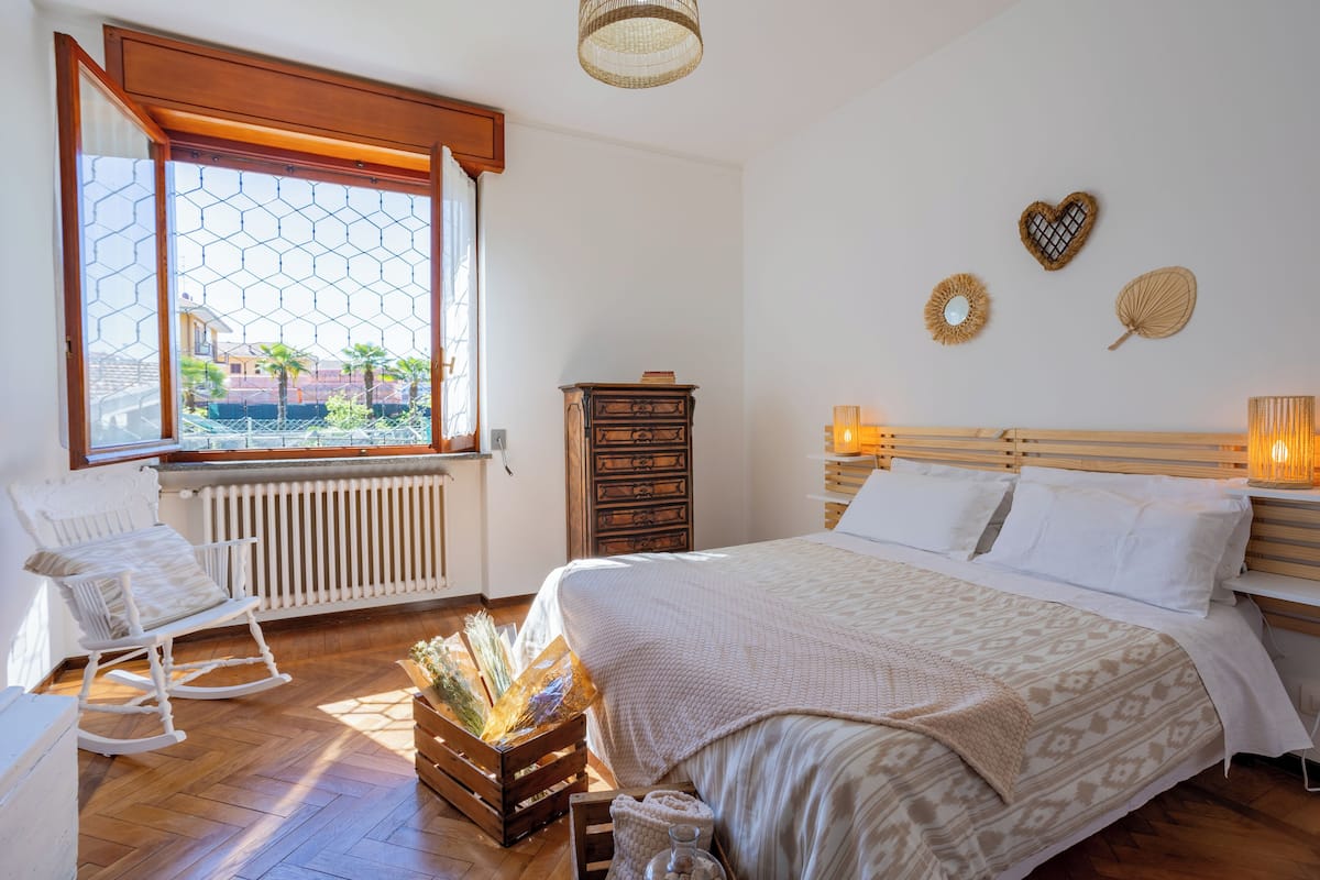 Une chambre à coucher avec un grand lit, un fauteuil à bascule, une commode en bois et une vue sur l’extérieur par une fenêtre à cadre en bois.