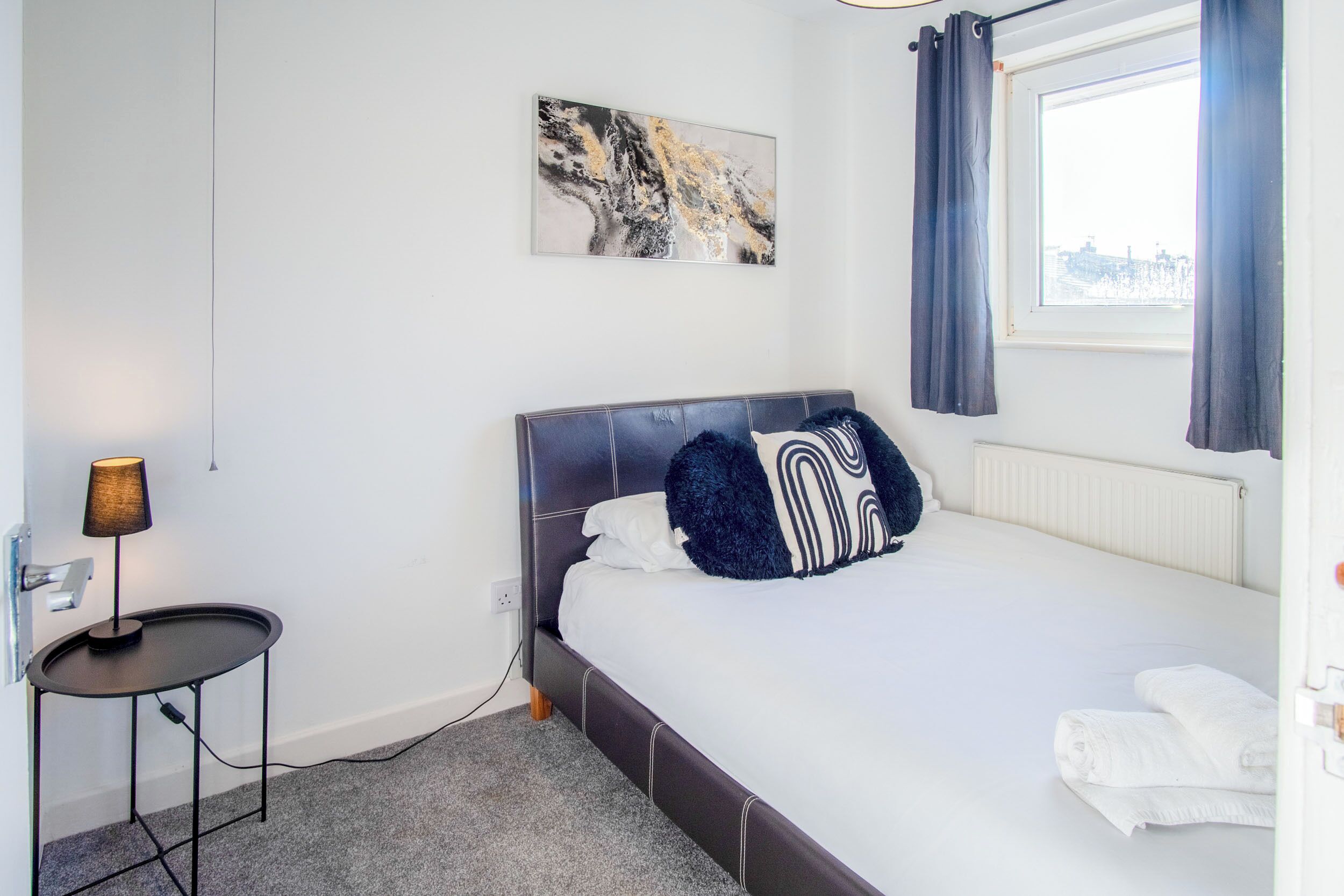 A bedroom with a bed, a bedside table with a lamp, a window with curtains, and a wall-mounted artwork.