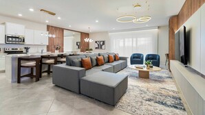 Una sala moderna con un sofá modular, una mesa de centro y un televisor. Se observa una cocina con microondas y armarios.